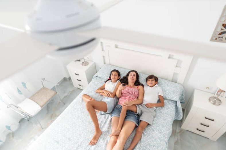 Guia Completo de Ventilador de Teto 2025 : Como Escolher o Modelo Ideal para Sua Casa 1 Família deitada na cama em um quarto claro e arejado, relaxando sob um ventilador de teto branco ligado.